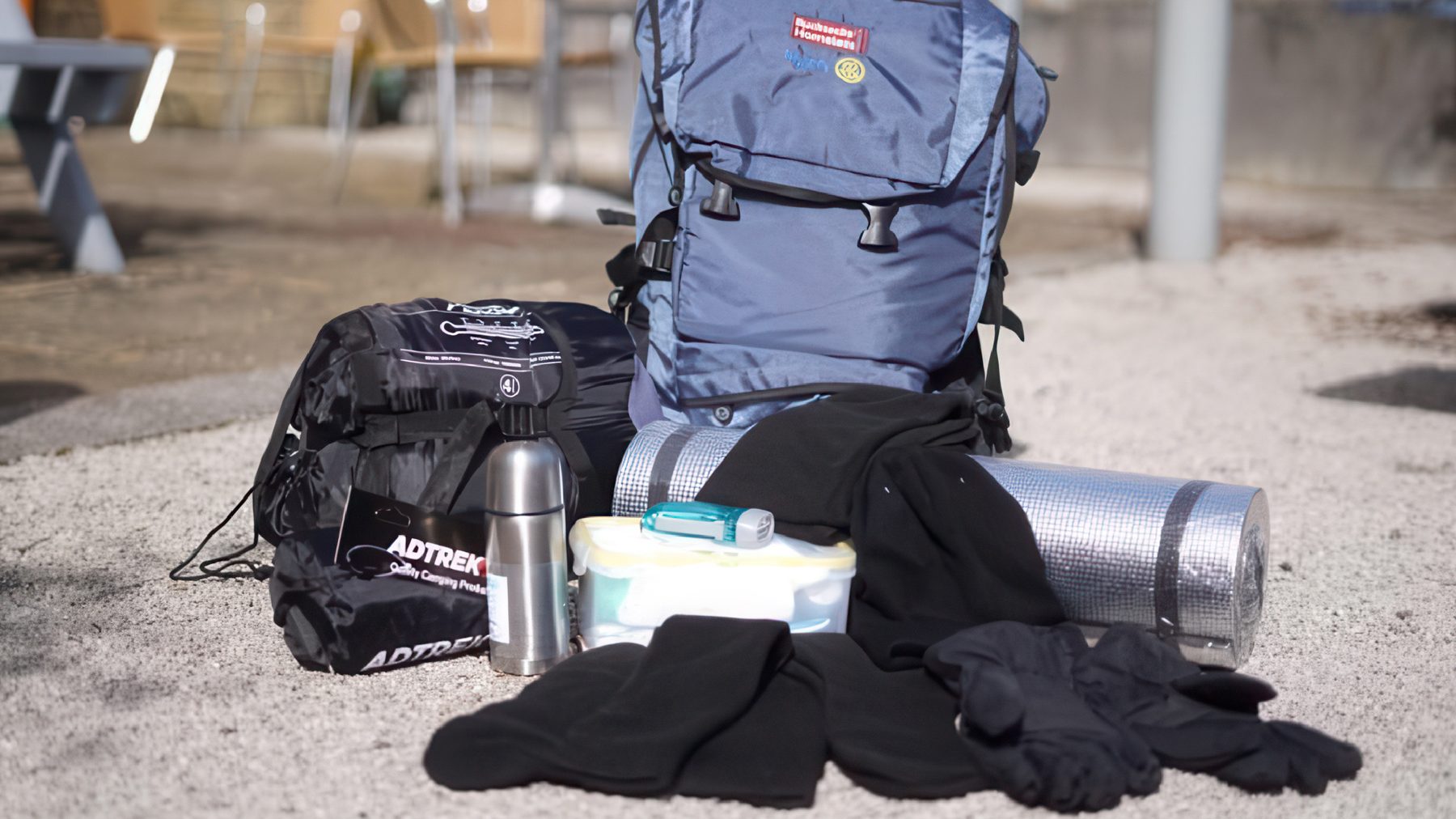 NEST1 Backpack, rolled sleeping pad, thermos, plastic container, gloves, and other camping gear on gravel