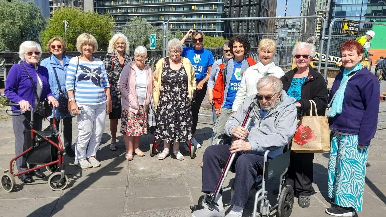 Group of older adults on a sunny city outing, smiling together with tall buildings behind them.