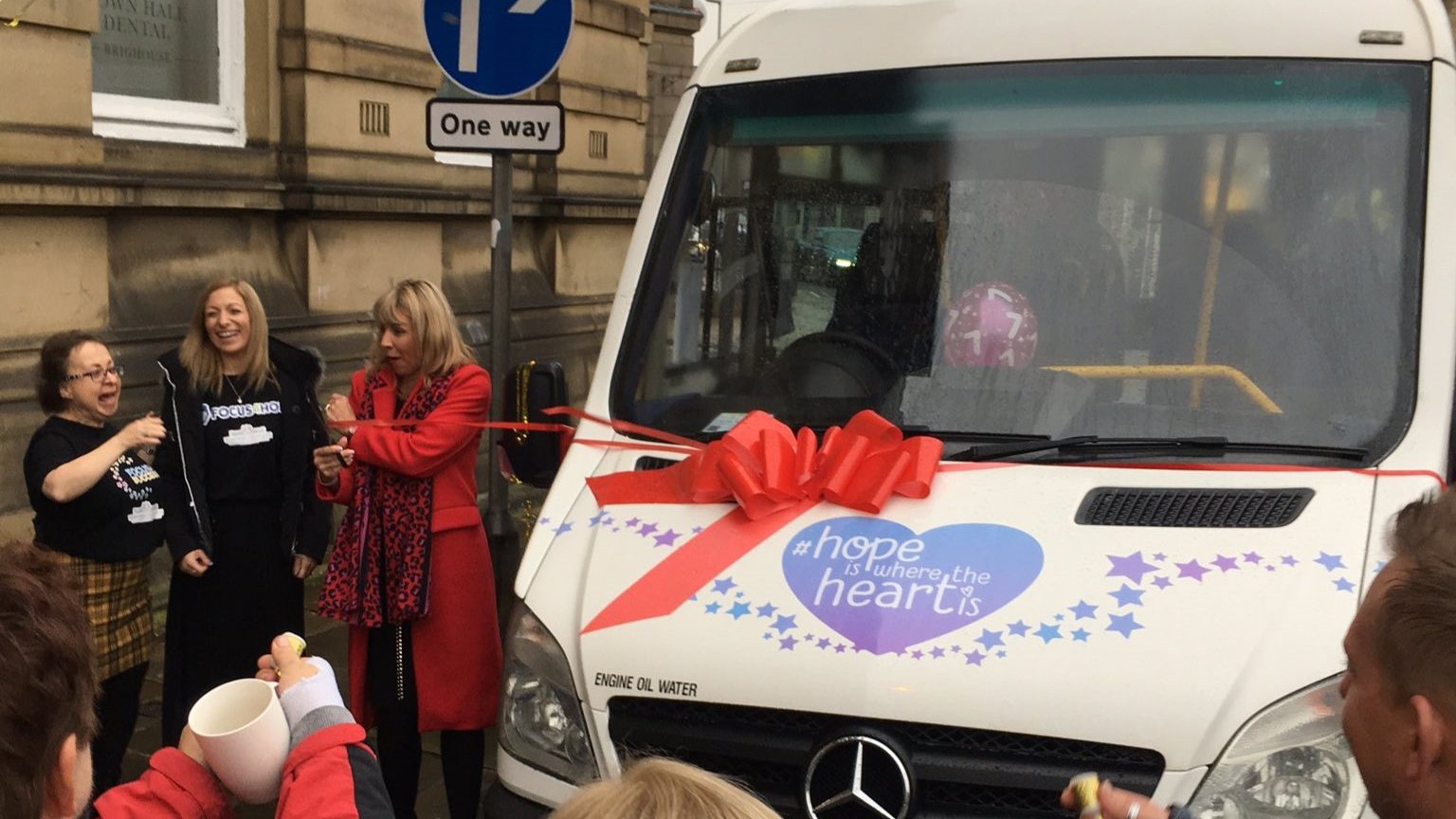 Group gathered on a street for a ribbon‑cutting ceremony beside a white van decorated with a red bow.