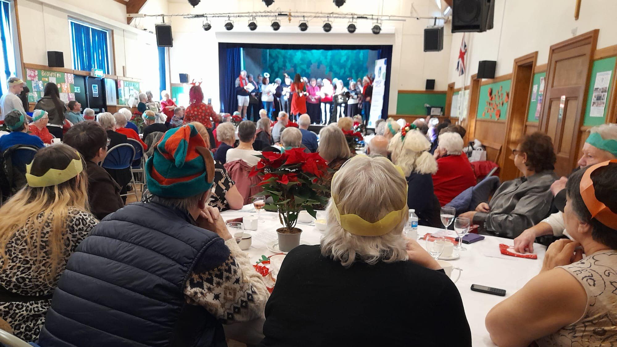 Community hall filled with older adults enjoying a Christmas party. Guests wear colourful paper crowns and festive hats while watching a group perform on stage. Tables are decorated with poinsettias and holiday items, creating a warm, cheerful atmosphere.